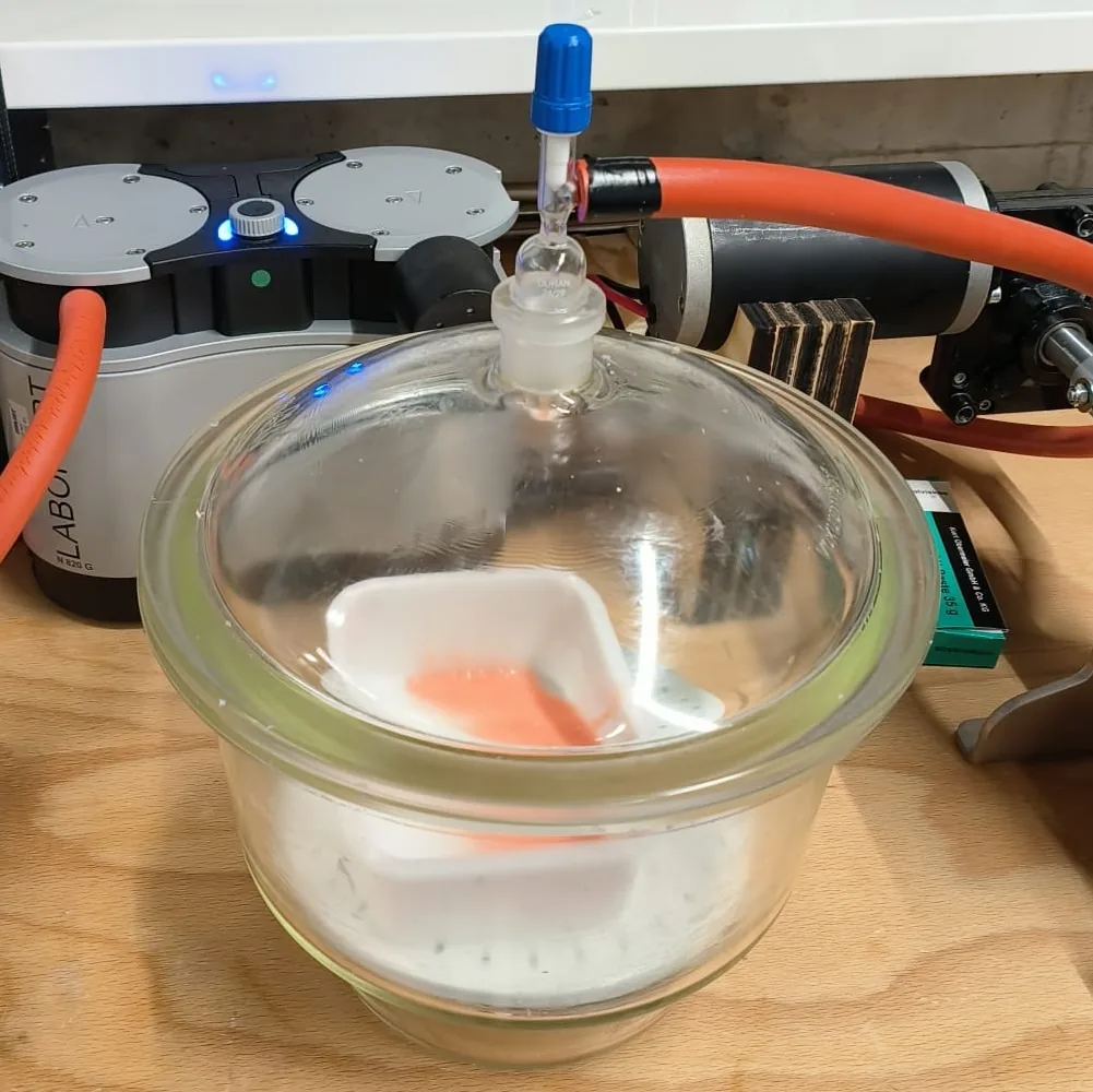 A glass vacuum desiccator on a bench, with a container of mixed silicone inside it and a vacuum pump behind.