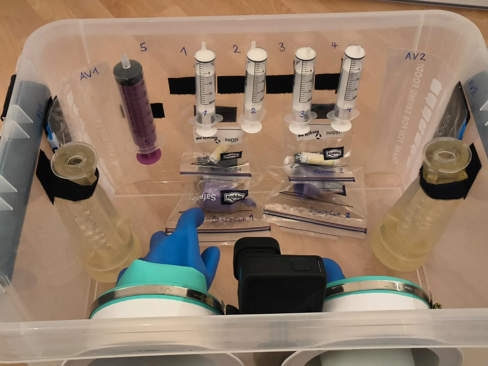 Inside the flight glovebox: two artificial vaginas labelled AV1 and AV2, numbered syringes, tampons and SpaceCups in labelled bags, and a camera, seen from above with a gloved hand reaching in.